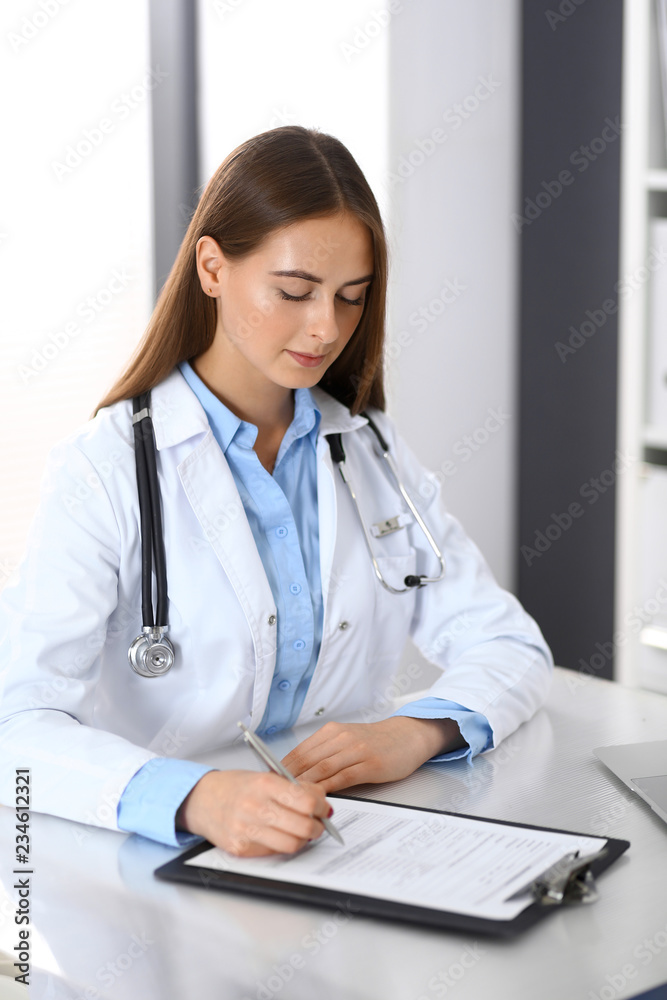 Doctor woman filling up medical form while sitting at the desk in hospital office. Physician at work. Medicine and health care concept Doctor woman filling up medical form while sitting at the desk in hospital office. Physician at work. Medicine and health care concept