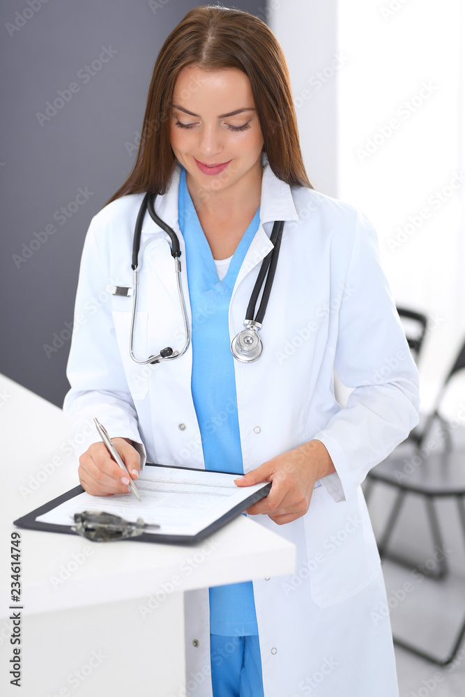 Doctor woman at work. Portrait of female physician filling up medical form while standing near reception desk at clinic or emergency hospital. Medicine and healthcare concept