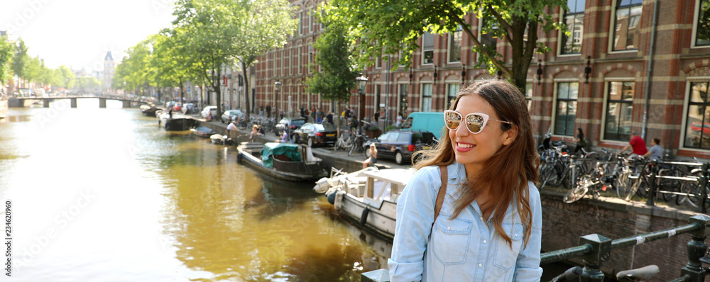Fototapeta premium Portret pięknej wesołej dziewczyny w okularach przeciwsłonecznych, patrząc z boku na jednym z typowych kanałów w Amsterdamie, Holandia. Panoramiczny widok banera.