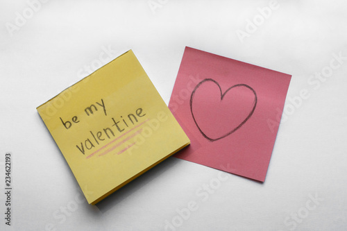 Be My Valentine handwritten inscription on the yellow sticker and heart on the pink paper, white background