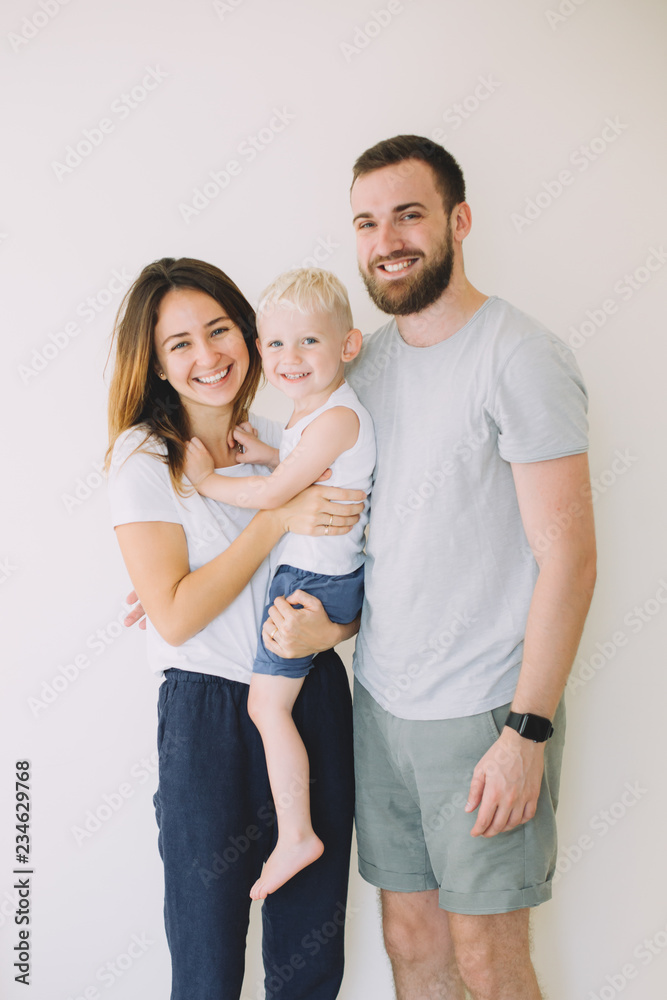 Happy young family with little son standing together in casuals near the empty wall - indoors - obrazy, fototapety, plakaty 