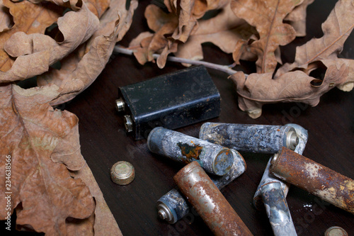Spent batteries, coated with corrosion. Different shapes and sizes. They lie on a dark background among the dead autumn leaves. Environmental protection, recycling.