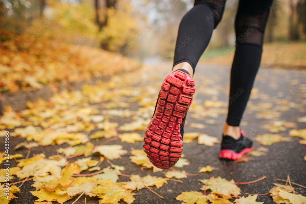 Picture of woman' legs in park on road. She runs. Woman wears black with red crosses and black sport pants. Yellow leaves are on road. Everything is covered with them.