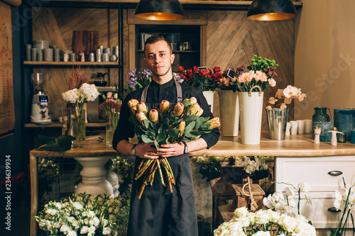 Portrait of male owner with lush bouquet of king Protea, assortment different flowers on wooden counter behind him. Man florist holding a flowers arrangements in modern interior floral butique photo