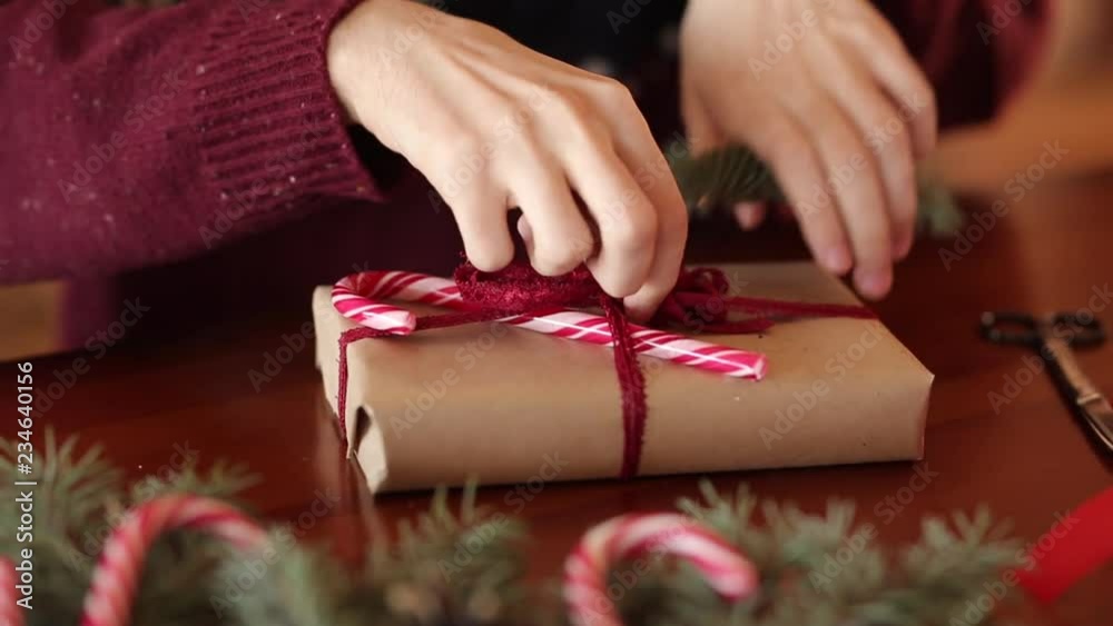 Close view of man's hands tying a bow on a gifts for Christmas. Male wrapping present boxes in paper adding fir branches, cones, cane candies on wooden table at home. Holidays preparations.