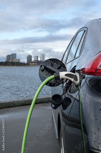Charging an electric car, with the city and water in the background, green charging cable and black car. copenhagen