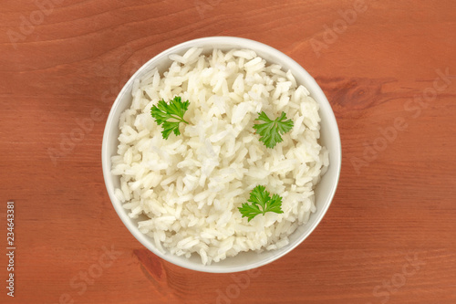 A bowl of cooked white jasmine rice, garnished with parsley leaves, shot from the top on a rustic wooden background with a place for text