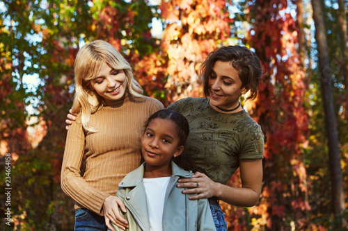 A beautiful couple of lesbian ladies posing with their adopted teenage daughter in the autumn park. The young women standing behind the kid, looking at her, smiling. The girl looking at the camera. photo