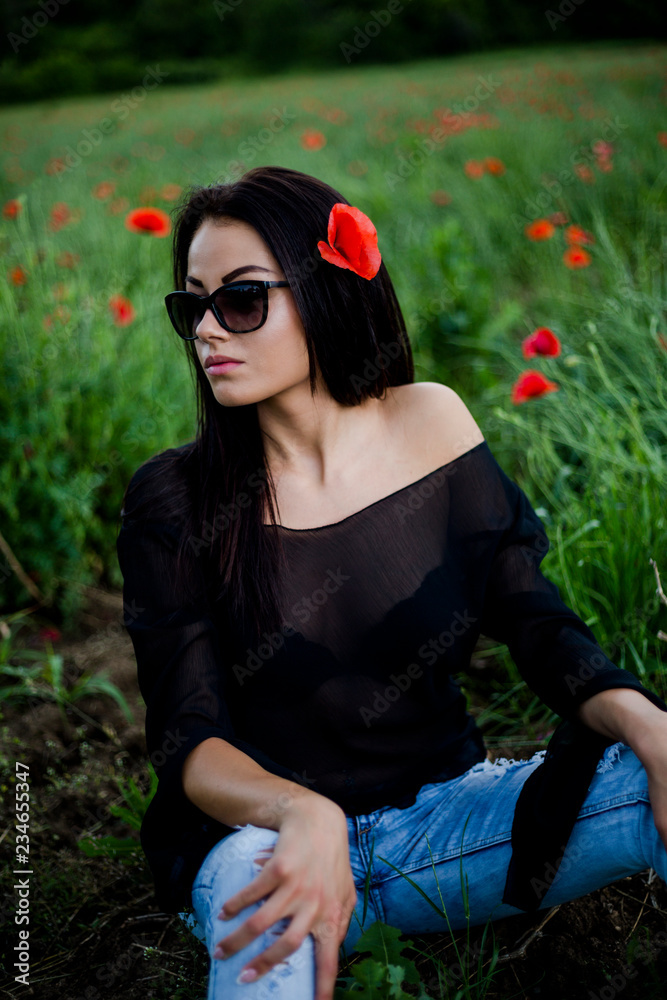 Beautiful brunette woman in sunglasses and stylish clothes sitting among on red poppies on meadow