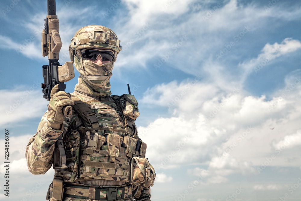 Half-length portrait of airsoft game player in army camouflage uniform,  tactical helmet, load carrier and face hidden behind mask, posing with  firearm replica in hands, cloudy sky on background foto de Stock