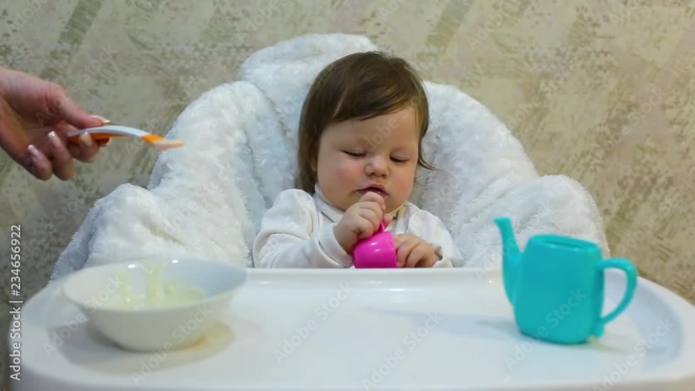 Cute toddler girl sitting in the child chair is refuses to fed porridge with spoon by mother hand Cute toddler girl sitting in the child chair is refuses to fed porridge with spoon by mother hand