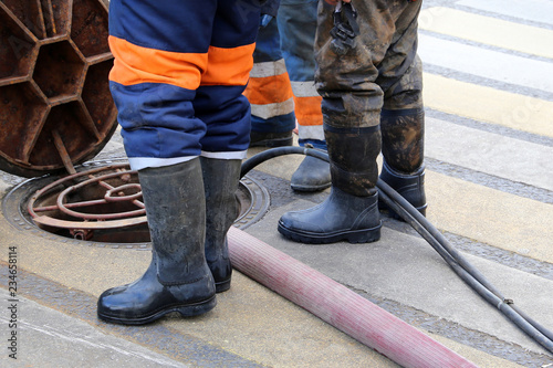 Workers in uniform and wellingtons stand over the open sewer hatch. Concept of repair of sewage, water supply system or underground utilities, cleaning drains, cable laying, water pipe accident