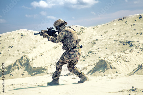 Man in military camouflage uniform with airsoft weapon bending down and running during armed conflict, military battle imitation in sandy or desert area. Airsoft player under fire searching cover