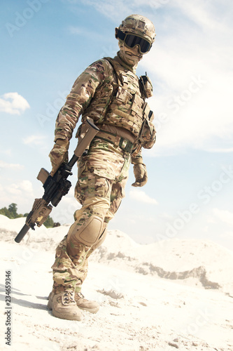 Airsoft player  military games participant in U.S. army infantry camouflage uniform  tactical mask  helmet and glasses  standing in desert area with combat service rifle or carbine replica in hands