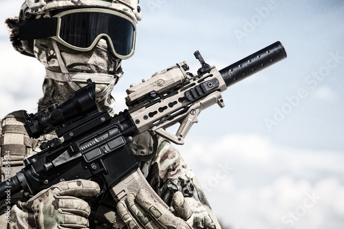 Half-length portrait of airsoft game player in army camouflage uniform, tactical helmet, load carrier and face hidden behind mask, posing with firearm replica in hands, cloudy sky on background