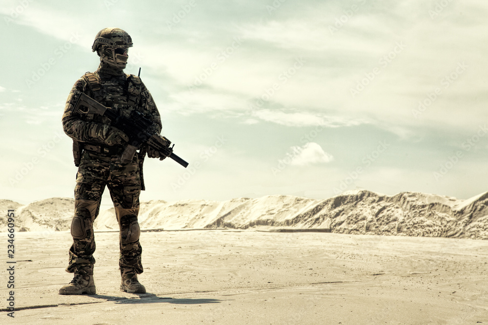 Special operations forces soldier in camouflage combat uniform, helmet and glasses walking in desert with service rifle in hands low angle view. War conflict, military campaign in Middle East region Special operations forces soldier in camouflage combat uniform, helmet and glasses walking in desert with service rifle in hands low angle view. War conflict, military campaign in Middle East region