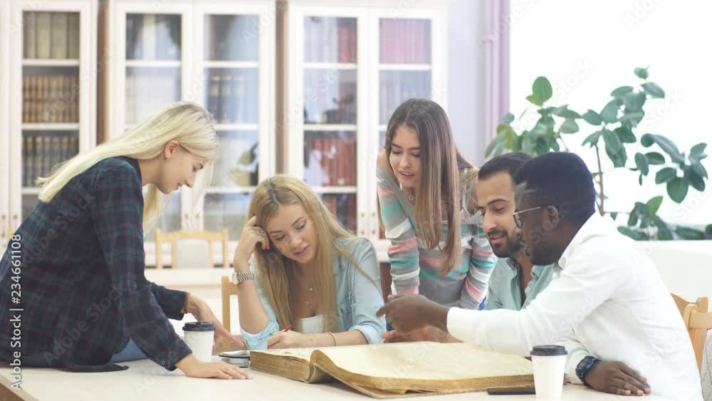 Multiracial students learning in college library, sitting at the table with big old reference textbook, writing down and discussing notes. Multiracial students learning in college library, sitting at the table with big old reference textbook, writing down and discussing notes.