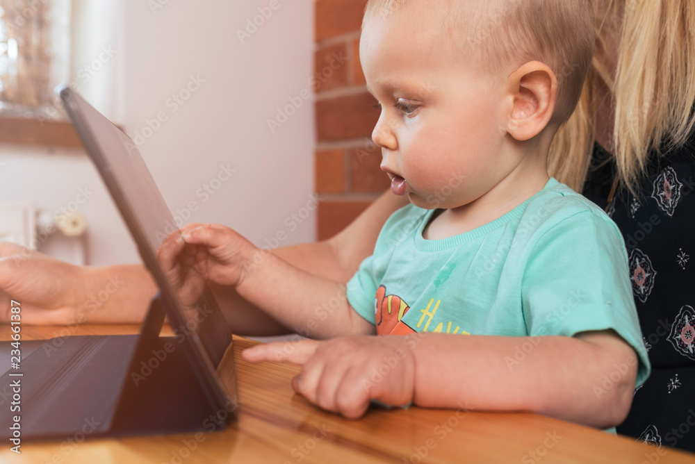 Mother and baby boy sitting at the table using digital tablet. Young mom teach little child how to use tablet with touchscreen watching a video or playing on digital tablet. Mother and son are playing Mother and baby boy sitting at the table using digital tablet. Young mom teach little child how to use tablet with touchscreen watching a video or playing on digital tablet. Mother and son are playing