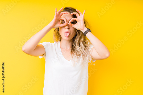 Young beautiful blonde woman over yellow background doing ok gesture like binoculars sticking tongue out, eyes looking through fingers. Crazy expression.