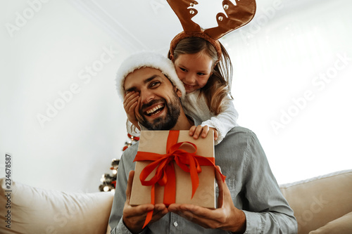 Christmas. Family. Love. Little girl is covering her dad's eyes making a surprise with a gift box. Near the Christmas tree at home