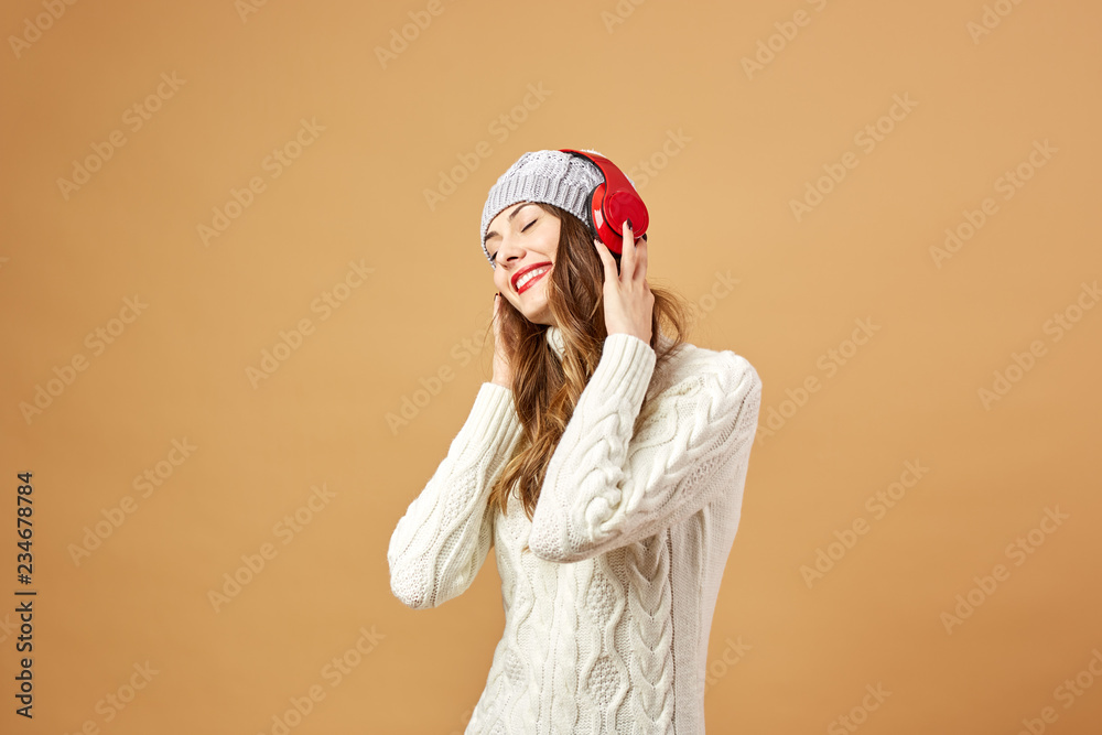 Smiling girl in red headphones dressed in white knitted sweater and hat stands on a beige background in the studio Smiling girl in red headphones dressed in white knitted sweater and hat stands on a beige background in the studio