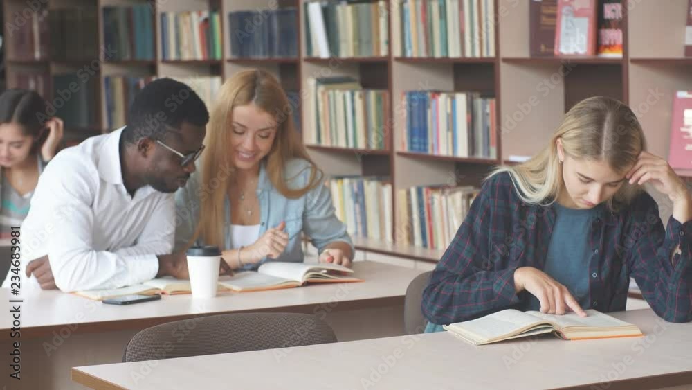 Smiling attractive blonde girl makes some notes while reading book in college library, looking at camera, sitting at desk with the other students on background Smiling attractive blonde girl makes some notes while reading book in college library, looking at camera, sitting at desk with the other students on background