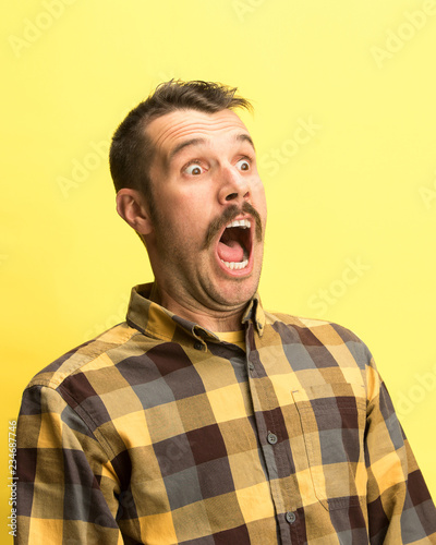 Fototapeta The surprised and astonished young man screaming with open mouth isolated on yellow background