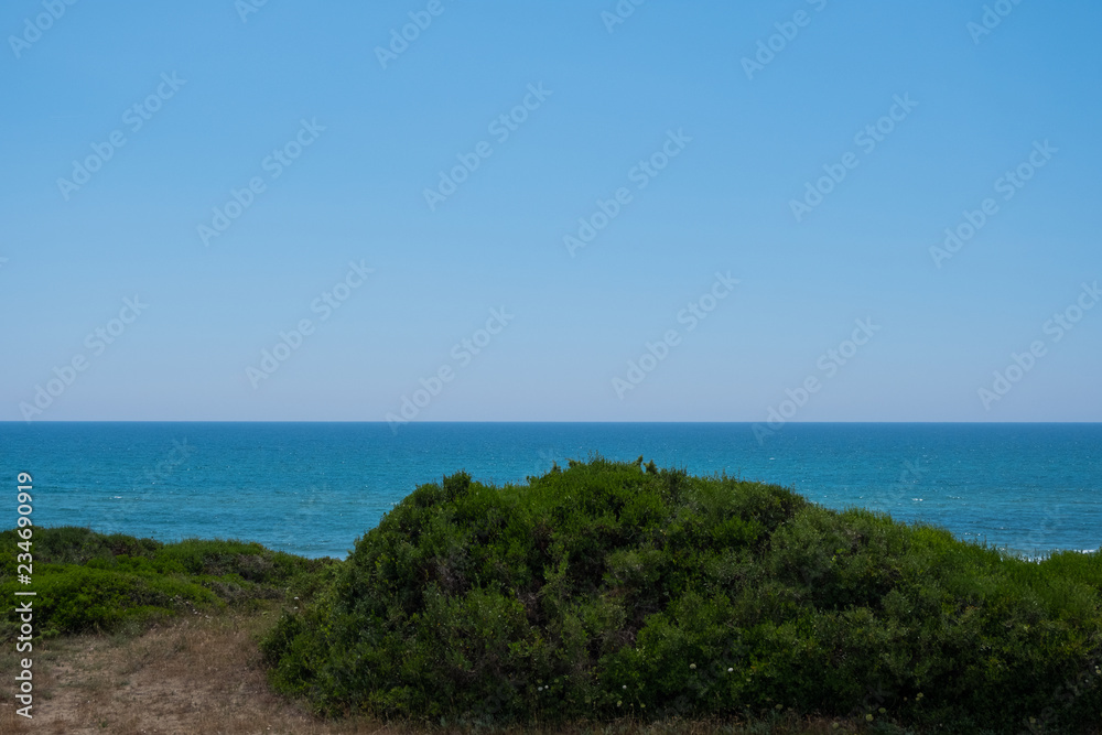 Blue sky and ocean water behind green hill.