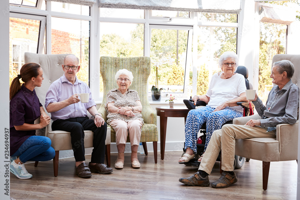 Male And Female Residents Sitting In Chairs And Talking With Carer In Lounge Of Retirement Home - obrazy, fototapety, plakaty 