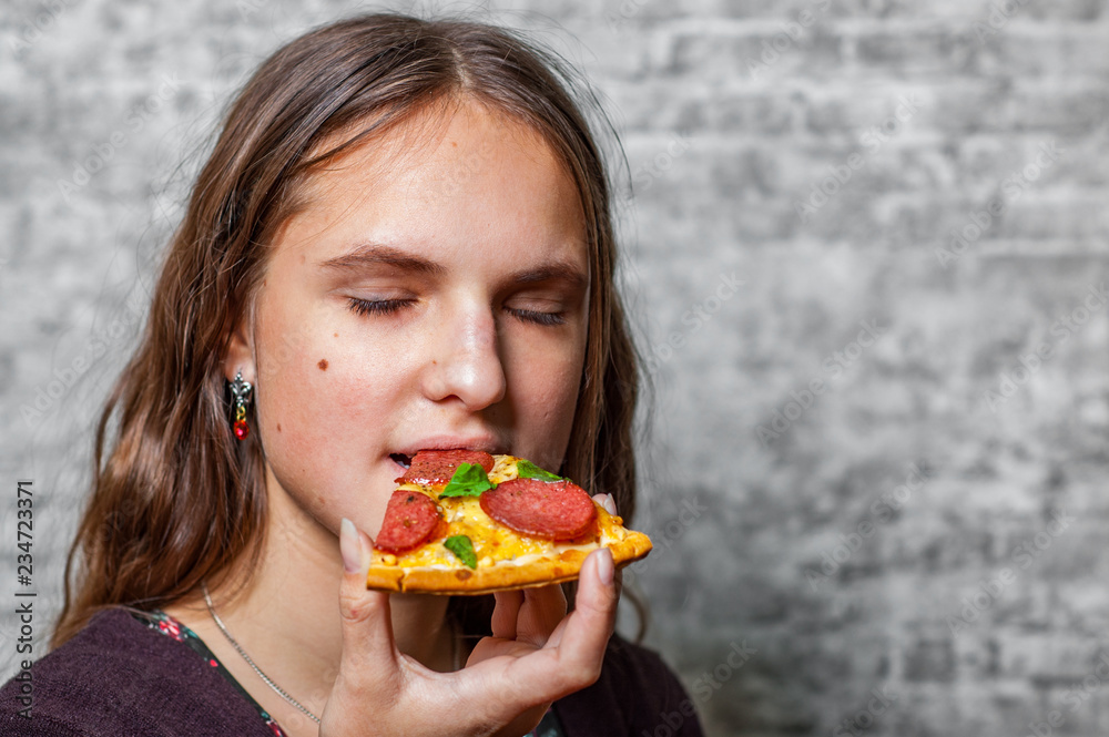 portrait of young teenager brunette girl with long hair eating slice of pizza on gray wall background