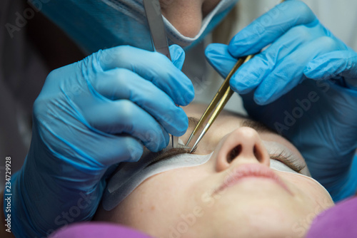 Eye lash extension procedure. Young beautiful woman eye with long false eye lashes. Close up macro shot of master making eyelashes in beauty salon.