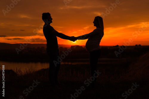 Silhouettes of husband and pregnant wife holding hand and enjoy spending time together outdoor.