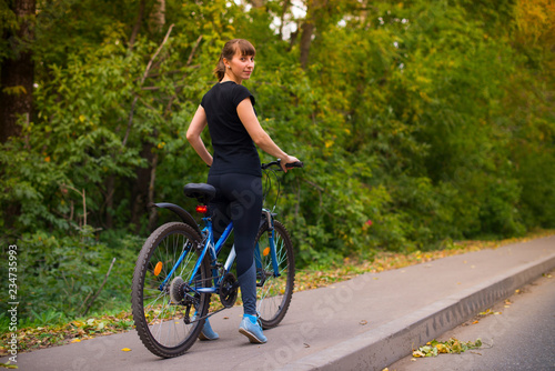 young beautiful athletic girl in sportswear on a blue bike stands half a turn. Trees on the background.