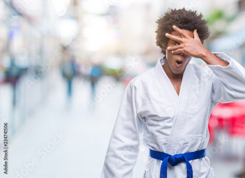 Afro american man wearing karate kimono over isolated background peeking in shock covering face and eyes with hand, looking through fingers with embarrassed expression.