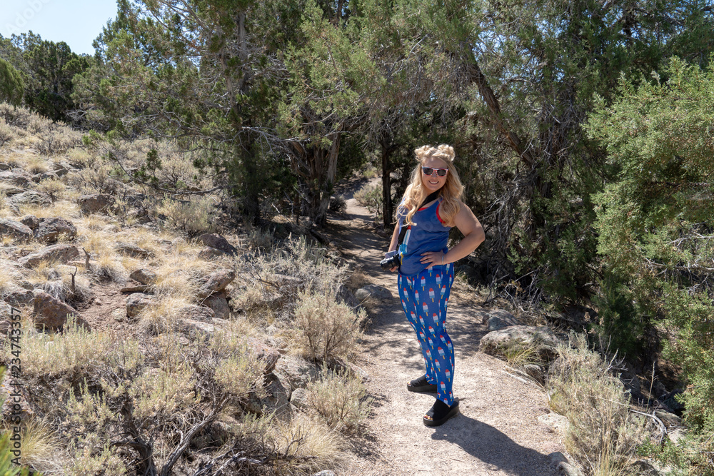 Young blond woman hiker wearing goofy American patriotic clothing and sunglasses on a dirt trail at the ghost town of Old Irontown, Utah