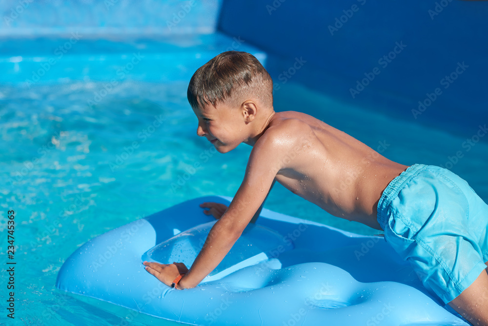 Smiling Caucasian boy swimming with inflatable mattress in pool at resort on family vacation.