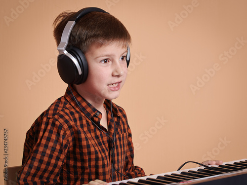 European boy is emotionally playing the electric piano. He is composing music, singing, and using the headphones. Portrait against peach background.