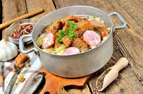 Garlic soup in an old saucepan with sausage, fresh garlic bulbs, cloves, colored pepper, wooden spoon, croutons and a towel on a rustic wooden table