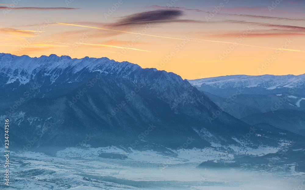 Idyllic winter landscape with snowy Piatra Craiului mountain range and misty valleys at sunset, Romania.