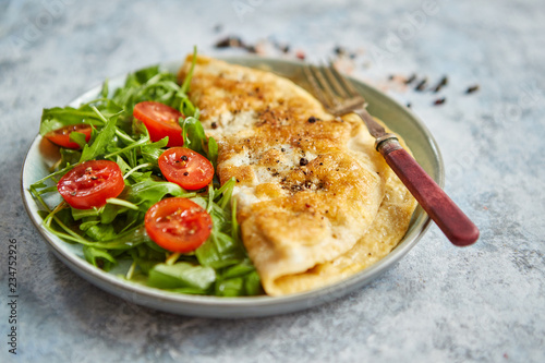 Classic egg omelette served with cherry tomato and arugula salad on side. Placed on white ceramic plate. Stone background with copy space. photo