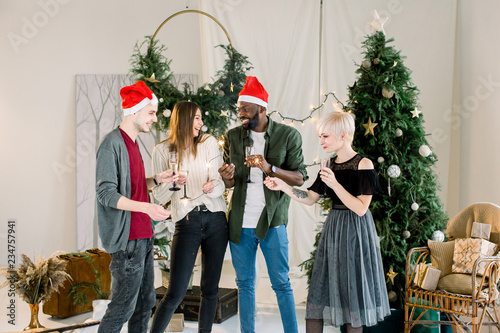 Merry Christmas and Happy New Year! Multiracial young creative people are celebrating holiday in cozy decorated studio. Group of young people are drinking champagne and holding bengal fires.