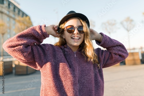 Outdoor portrait of a smiling teen girl 13, 14 years old in a hat and sunglasses. City background, golden hour. photo