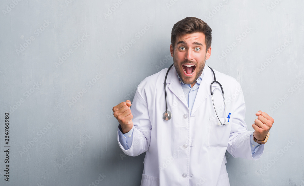 Handsome young doctor man over grey grunge wall celebrating surprised and amazed for success with arms raised and open eyes. Winner concept. Handsome young doctor man over grey grunge wall celebrating surprised and amazed for success with arms raised and open eyes. Winner concept.