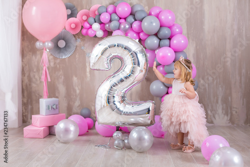 Princess baby girl celebrating life event wearing golden crown and pink airy dress. Cute girl posing in pastel colors studio shoot with air baloons and cake.