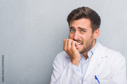 Handsome young professional man over grey grunge wall wearing white coat looking stressed and nervous with hands on mouth biting nails. Anxiety problem.