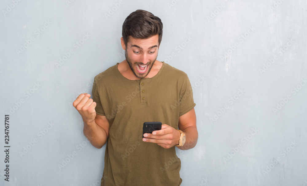 Handsome young man over grey grunge wall texting a message using smartphone screaming proud and celebrating victory and success very excited, cheering emotion