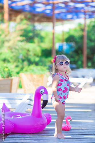 Baby girl on beach with pink flamingo dressed in stylish swimming wear posing on wooden floor. Scene on vacation on sea side of a cute lady smiling and enjoy life time childhood infancy.