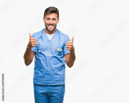 Young handsome doctor surgeon man over isolated background success sign doing positive gesture with hand, thumbs up smiling and happy. Looking at the camera with cheerful expression, winner gesture.
