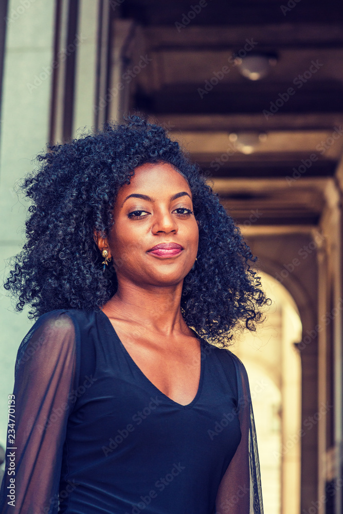 Portrait of Young African American Woman in New York. Young black female  college student with afro hairstyle wearing black shirt, standing on narrow  vintage street on campus, looking at you.. Stock Photo