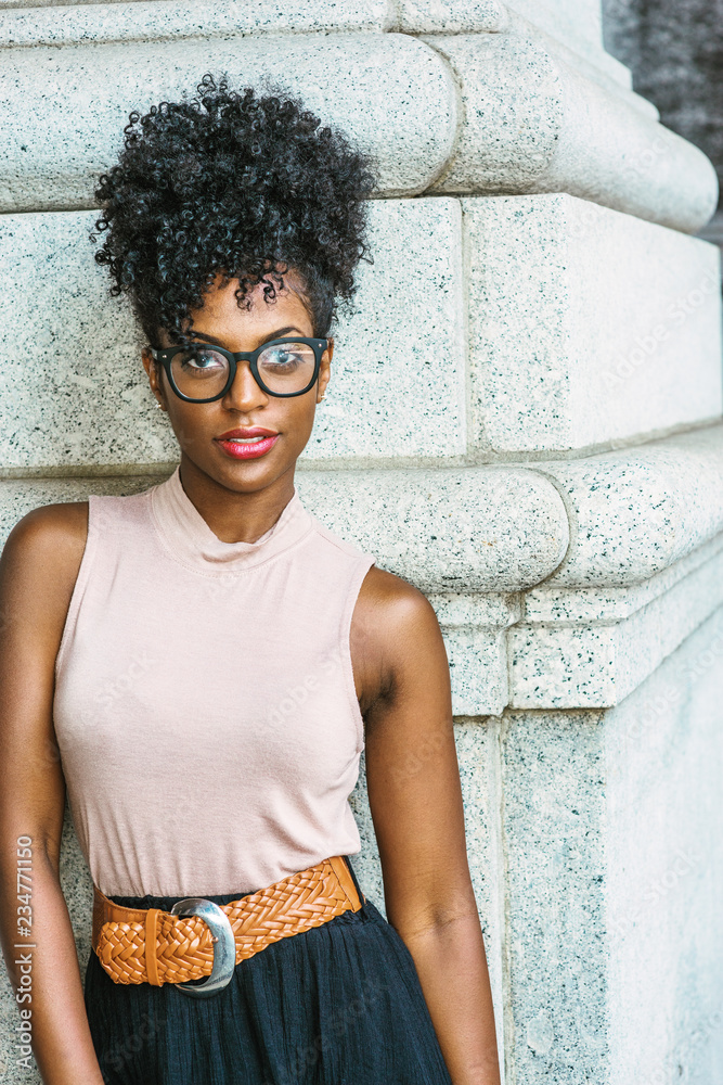 Portrait of Young African American Woman in New York. Young black female  college student with afro hairstyle wearing sleeveless light color top,  black skirt, eye glasses, standing by column on street Stock